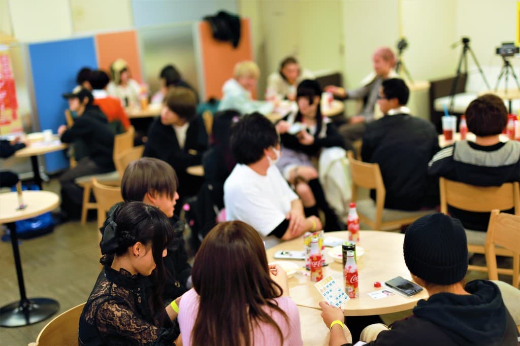 明るい会場で円卓を囲むメンバーと三脚の配信カメラ。交流イベントでの様子